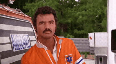 A Man With A Mustache Is Standing In Front Of An Ambulance And Looking At The Camera . GIF