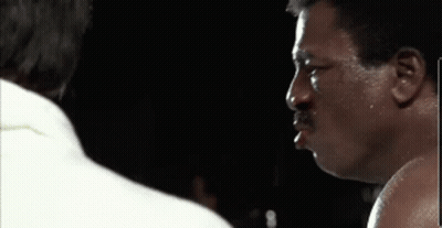 A Man With A Mustache Is Standing Next To Another Man In A Boxing Ring In A Dark Room . GIF