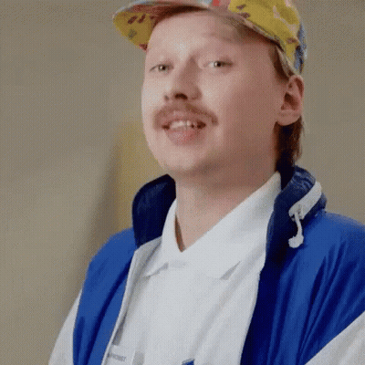 A Man With A Mustache Is Wearing A Blue Jacket And A Hat . GIF