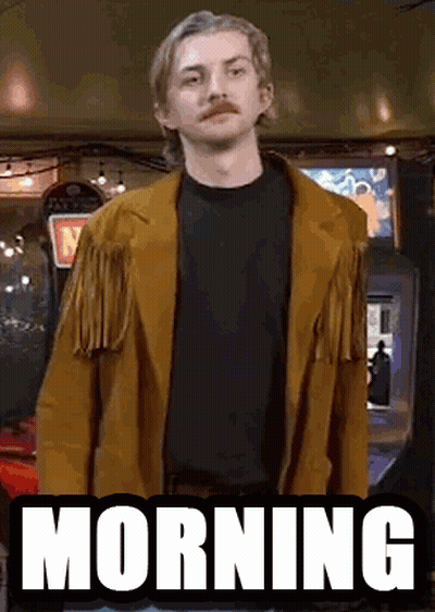 A Man With A Mustache Is Wearing A Brown Jacket And Standing In Front Of An Arcade Machine That Says Morning . GIF