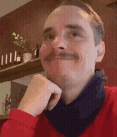 A Man With A Mustache Is Wearing A Red Sweater And A Black Scarf Around His Neck . GIF
