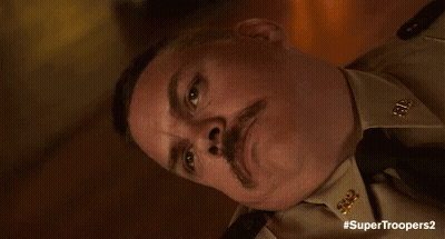 A Man With A Mustache Is Wearing A Tan Uniform And Tie And Has The Hashtag #Supertroopers2 GIF