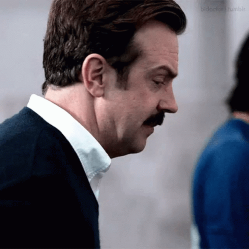 A Man With A Mustache Is Wearing A White Shirt And A Blue Sweater . GIF