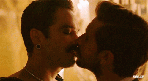 A Man With A Mustache Kisses Another Man With A Necklace Around His Neck GIF