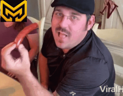A Man With A Mustache Wearing A Black Shirt And A Black Hat Is Eating A Hot Dog GIF