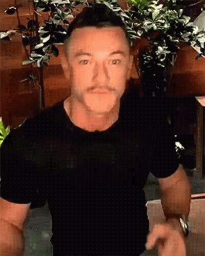 A Man With A Mustache Wearing A Black T-Shirt Is Standing In Front Of A Plant . GIF