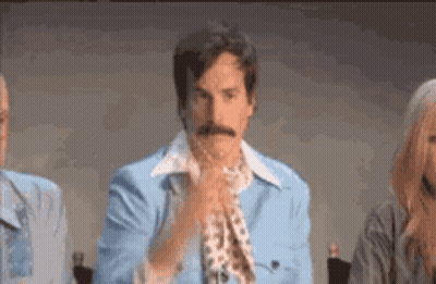 A Man With A Mustache Wearing A Blue Jacket And Tie Is Sitting In Front Of A Microphone . GIF