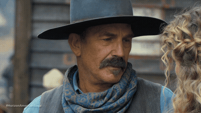A Man With A Mustache Wearing A Cowboy Hat And Scarf Talks To A Woman In A Horizon American Saga Ad GIF