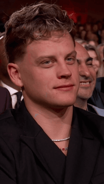 A Man With A Necklace Around His Neck Is Sitting In A Crowd Of People GIF