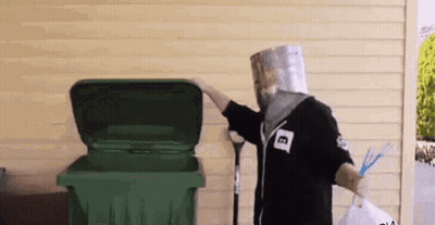 A Man With A Paper Bag On His Head Is Standing Next To A Green Trash Can . GIF