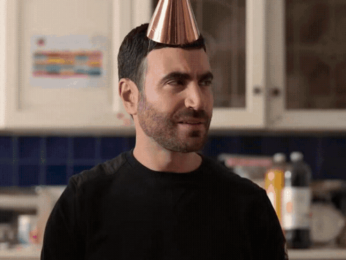 A Man With A Party Hat On His Head Looks At The Camera GIF