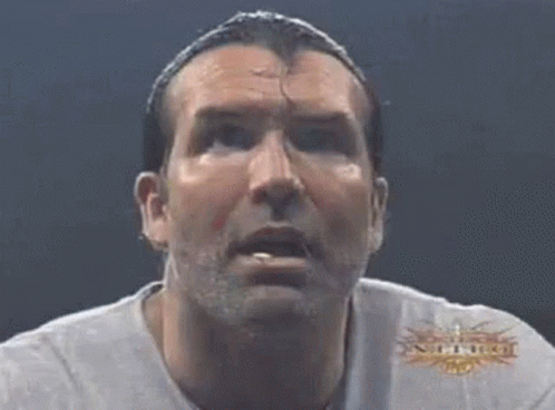 A Man With A Ponytail And A White Shirt Is Looking At The Camera . GIF