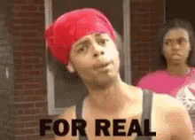 A Man With A Red Bandana On His Head Is Standing In Front Of A Brick Building . GIF