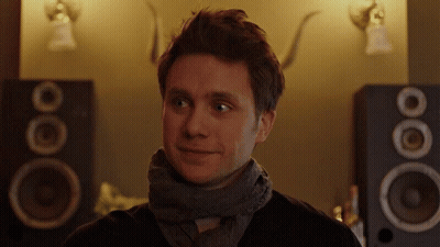 A Man With A Scarf Around His Neck Is Smiling In Front Of A Pair Of Speakers GIF