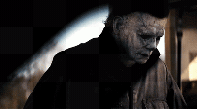 A Man With A Scary Mask On His Face Is Standing In A Dark Room . GIF