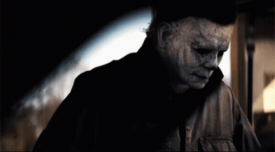 A Man With A Scary Mask On His Face Is Standing In A Dark Room . GIF