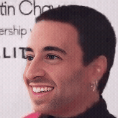 A Man With A Shaved Head And Earrings Is Smiling In Front Of A Sign That Says Tin Chow . GIF