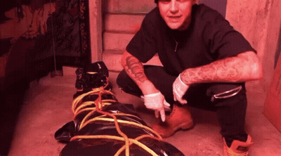 A Man With A Tattoo On His Arm Is Kneeling Next To A Black Bag Tied With Rope GIF