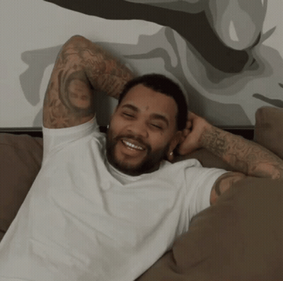 A Man With A Tattoo On His Arm Is Laying On A Couch And Smiling GIF