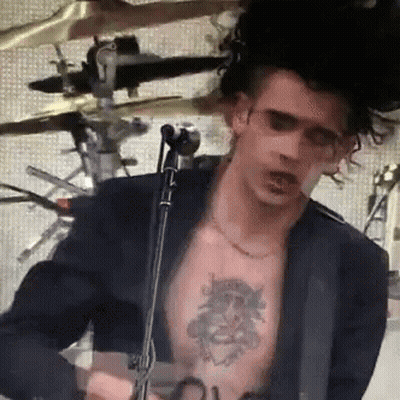 A Man With A Tattoo On His Chest Is Playing A Guitar In Front Of A Microphone GIF