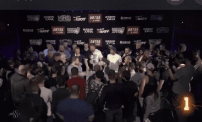 A Man With A Tattoo On His Face Stands In A Crowd With A Security Guard Behind Him GIF