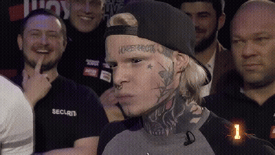 A Man With A Tattoo On His Face Stands In Front Of A Crowd Of People Wearing Security Shirts GIF