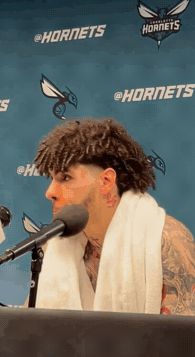 A Man With A Towel Around His Neck Is Speaking Into A Microphone In Front Of A Hornets Banner GIF