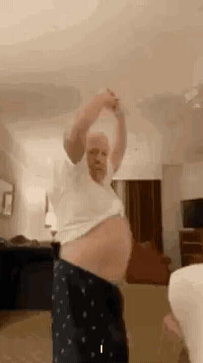 A Man With A Very Large Belly Is Dancing In A Living Room With A Woman . GIF