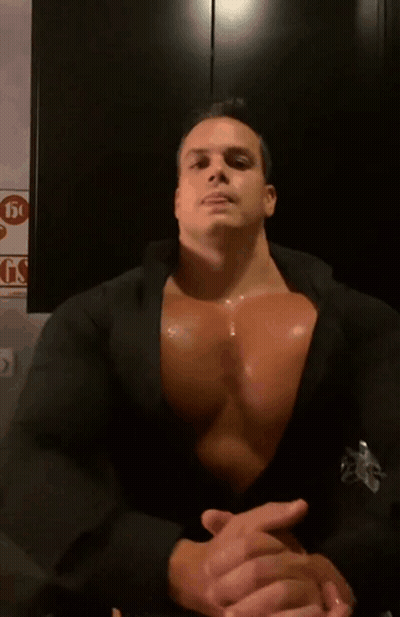 A Man With A Very Large Chest Is Sitting In Front Of A Sign That Says Gs GIF