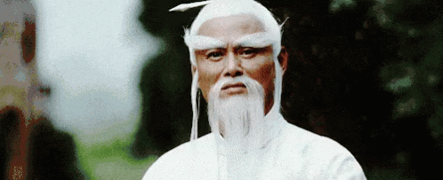 A Man With A White Beard And White Hair Is Standing In Front Of A Tree And Looking At The Camera . GIF