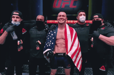 A Man With An American Flag Wrapped Around His Shoulders Stands In Front Of A Sign That Says Fc GIF