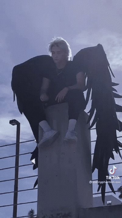 A Man With Angel Wings Is Sitting On A Concrete Wall GIF
