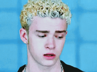A Man With Blonde Curly Hair Is Wearing A Black Shirt And A Chain Around His Neck . GIF