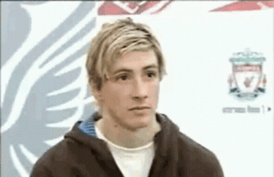 A Man With Blonde Hair And A Brown Jacket Is Standing In Front Of A Liverpool Logo . GIF