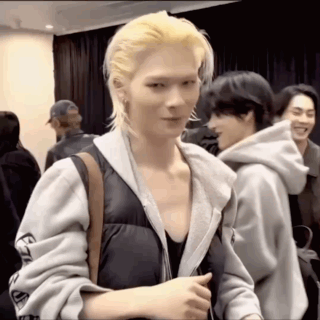 A Man With Blonde Hair And A Hoodie Is Standing In A Room With Other People . GIF