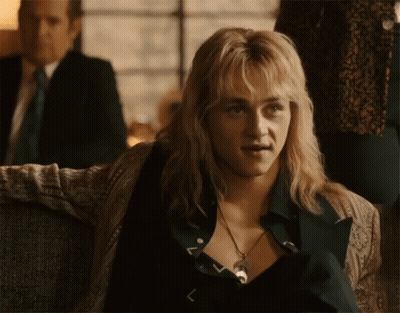 A Man With Blonde Hair And A Necklace Is Sitting On A Couch In Front Of A Man In A Suit . GIF