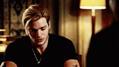 A Man With Blonde Hair And A Tattoo On His Arm Is Sitting At A Table With A Lamp In The Background . GIF