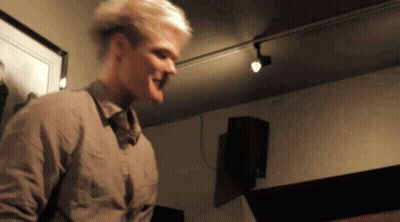 A Man With Blonde Hair Is Standing In A Room With A Picture On The Wall Behind Him GIF