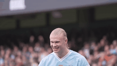 A Man With Blonde Hair Is Wearing A Blue Shirt And Smiling On A Soccer Field . GIF
