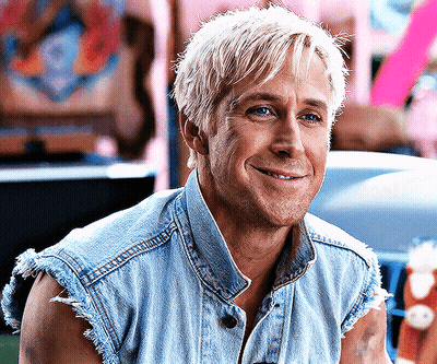 A Man With Blonde Hair Wearing A Denim Vest Is Smiling GIF