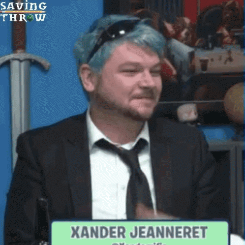 A Man With Blue Hair Is Sitting In Front Of A Sign That Says Xander Jeanneret GIF