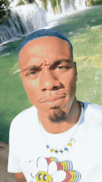 A Man With Blue Hair Is Wearing A White Shirt With A Bee On It In Front Of A Waterfall GIF