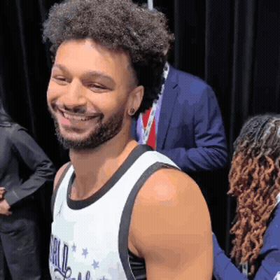 A Man With Curly Hair And A Beard Is Smiling For The Camera While Wearing A Basketball Jersey . GIF
