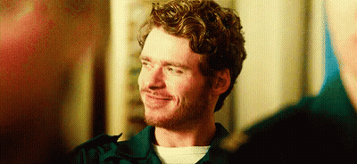 A Man With Curly Hair And A Beard Is Smiling While Standing Next To Another Man . GIF