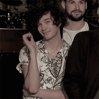 A Man With Curly Hair And A Beard Is Standing Next To Another Man In A Room . GIF