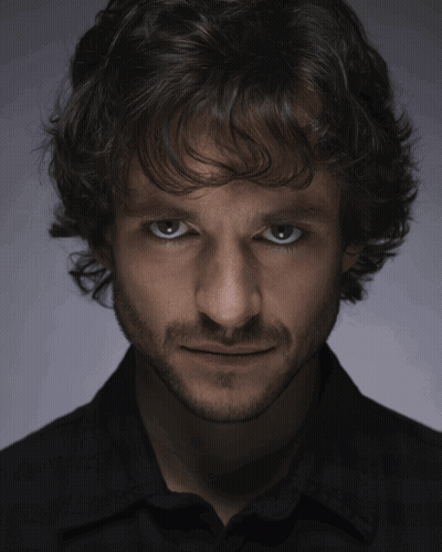 A Man With Curly Hair And A Beard Looks At The Camera With A Serious Look On His Face GIF