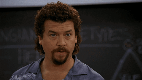 A Man With Curly Hair And A Beard Stands In Front Of A Blackboard That Says ' Ncaa ' On It GIF