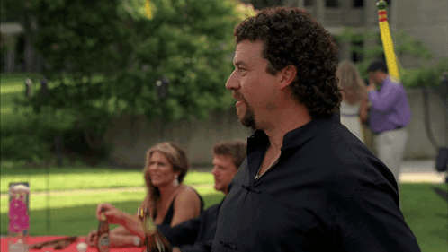 A Man With Curly Hair And A Beard Stands In Front Of A Table GIF