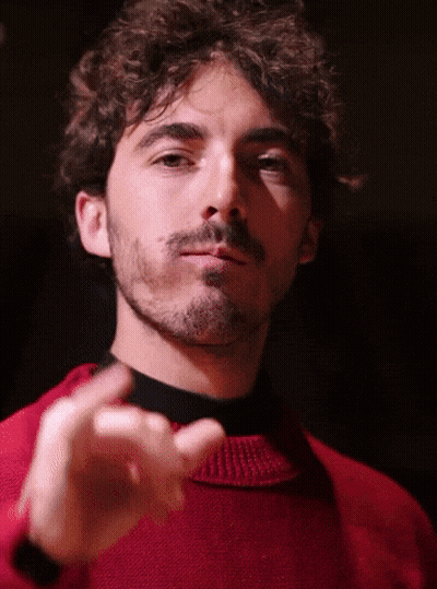 A Man With Curly Hair And A Beard Wears A Red Sweater And Black Turtleneck GIF