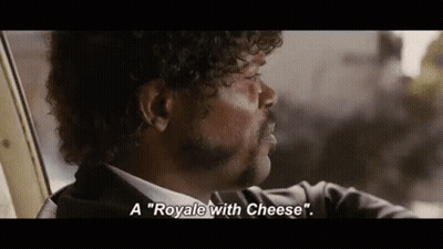 A Man With Curly Hair And A Mustache Is Sitting In A Car And Talking About A Royale With Cheese . GIF
